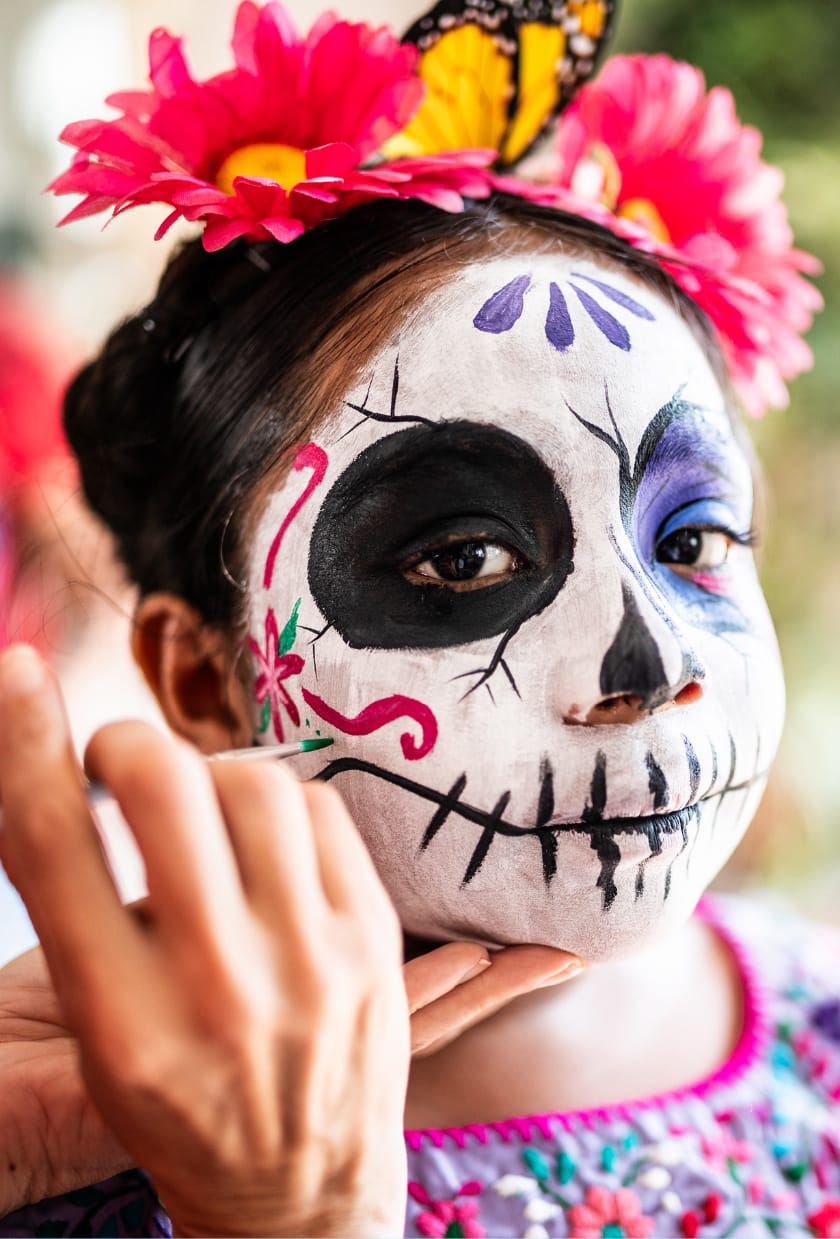 Une jeune fille se fait maquiller le visage en blanc pour ressembler à un squelette. Il y a des lignes noires à la bouche. Un oeil est noir et l’autre bleu. Il y a aussi des décorations de fleurs.